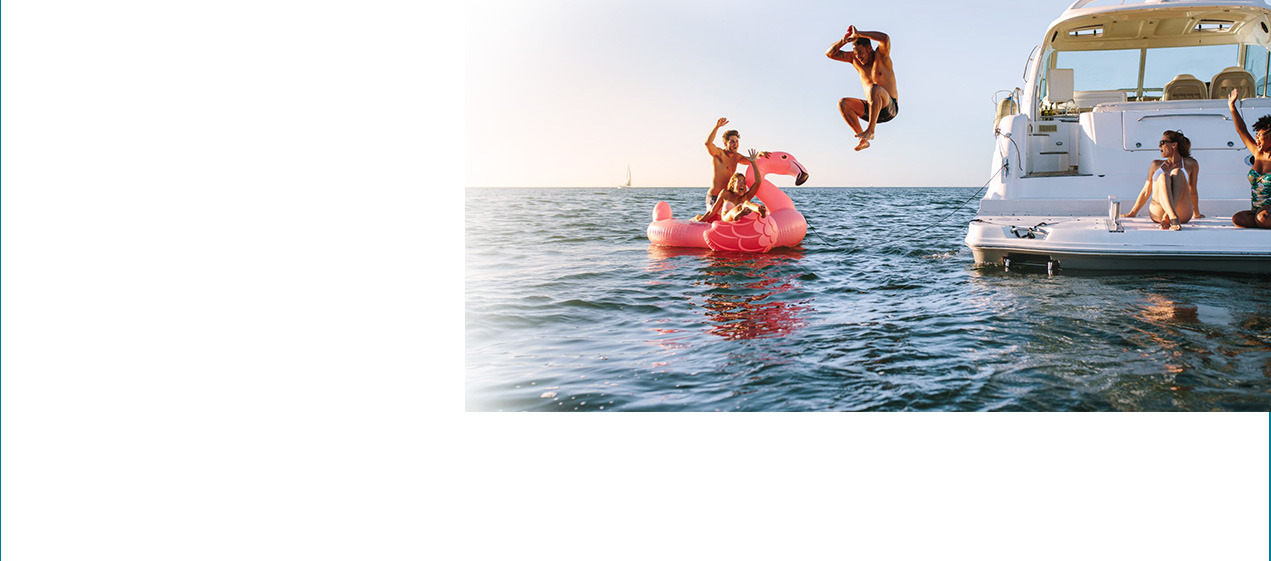 There’s a boat floating in lake with people relaxing on the back of the boat. A young man is jumping off the boat into the water to splash his friends who are floating on a nearby pink flamingo float. 