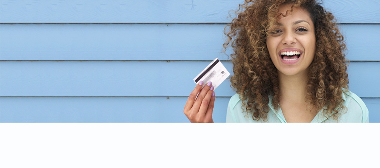 There’s a young lady standing in front of a blue wall holding a debit card in her hand and smiling