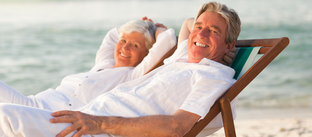 Retired couple dressed in all white is sitting in beach chairs smiling with the ocean behind them.